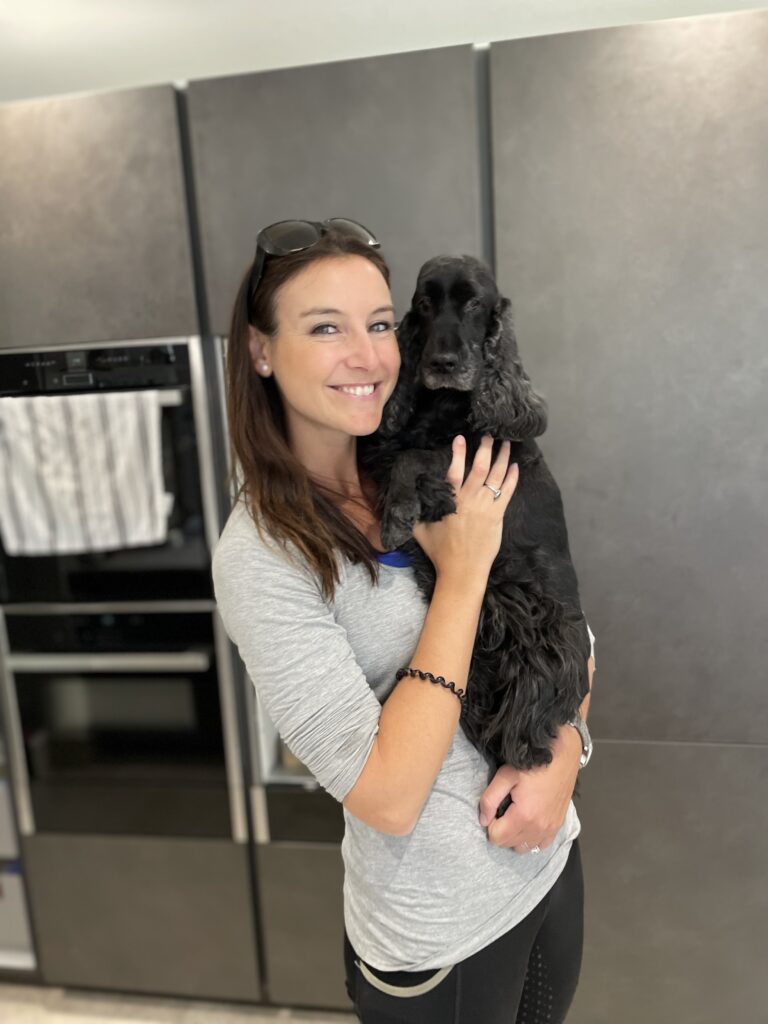 Ellie Kelland, owner of BARF time, smiling while holding in her arms her late black English Cocker Spaniel Clo. Clo has some white hair around her nose, showing her old age. Ellie is wearing a light grey long sleeved top and black trousers. They are standing in a kitchen with dark grey cupboards in the background. The scene conveys warmth and love.