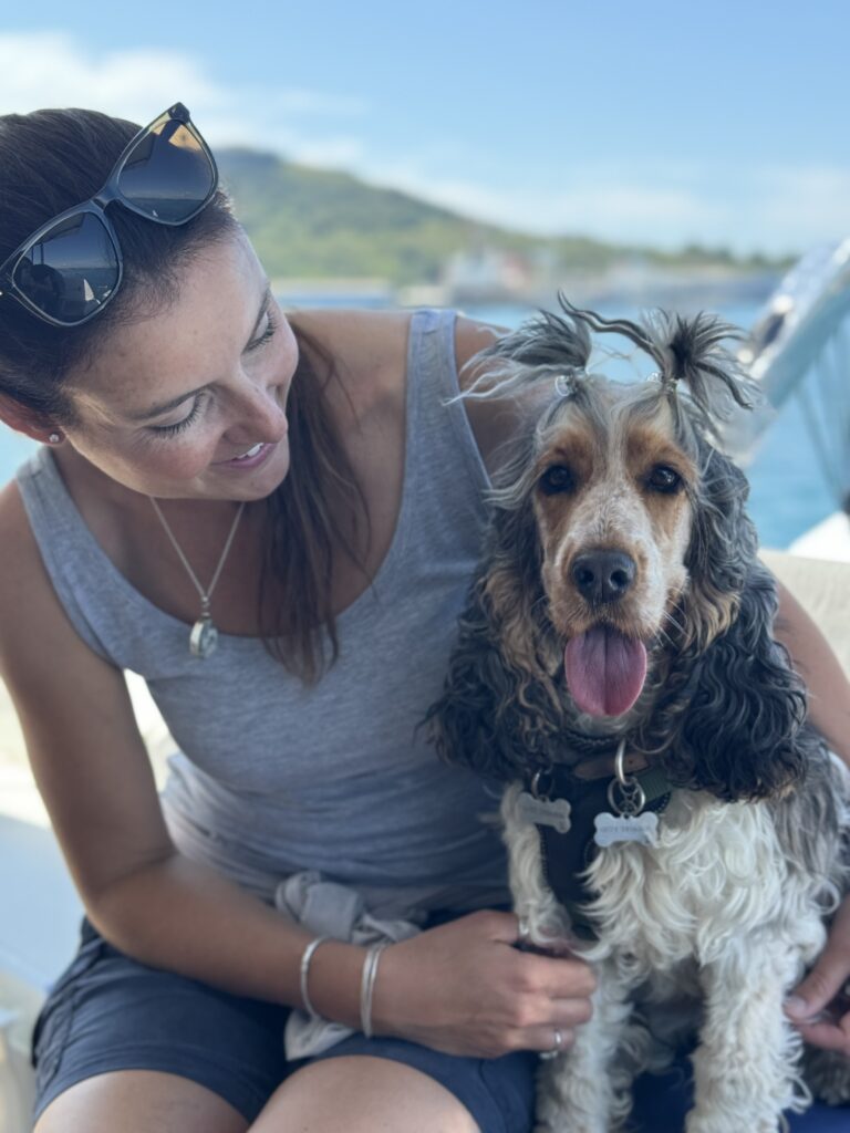 Ellie Kelland, owner of BARF time, is sitting down and smiling at her grey eared, brown faced and white legs, rescue English Cocker Spaniel girl Brandy, who is sitting next to her and has her head hair up in two cute small bunches. Brandy is also smiling with her tongue out. There is a blurred sea view in the background on a sunny day. The scene conveys warmth, love and happiness.